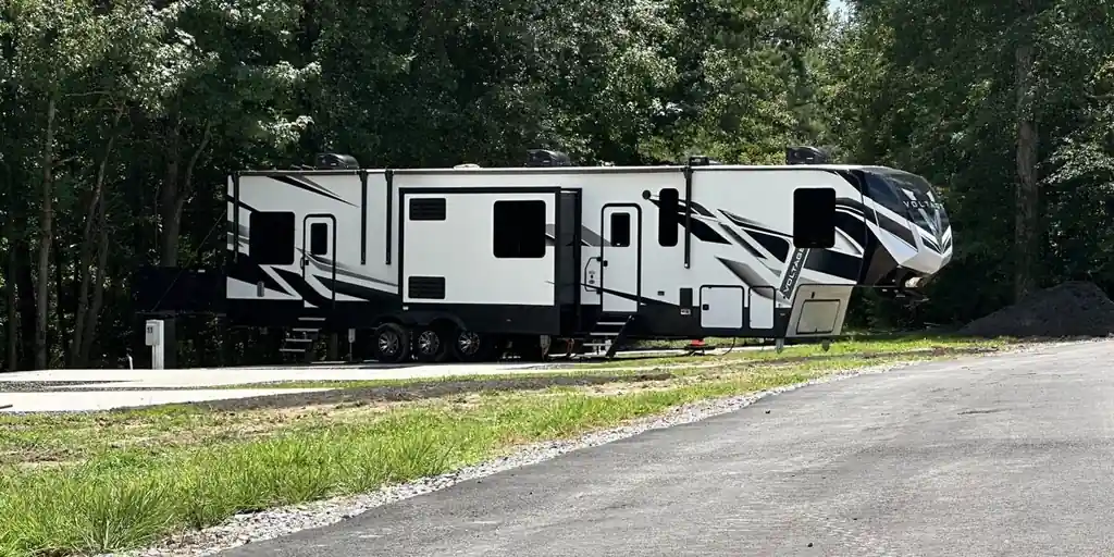 A large fifth-wheel parked on a pad edged by the asphalt loop and hardwoods